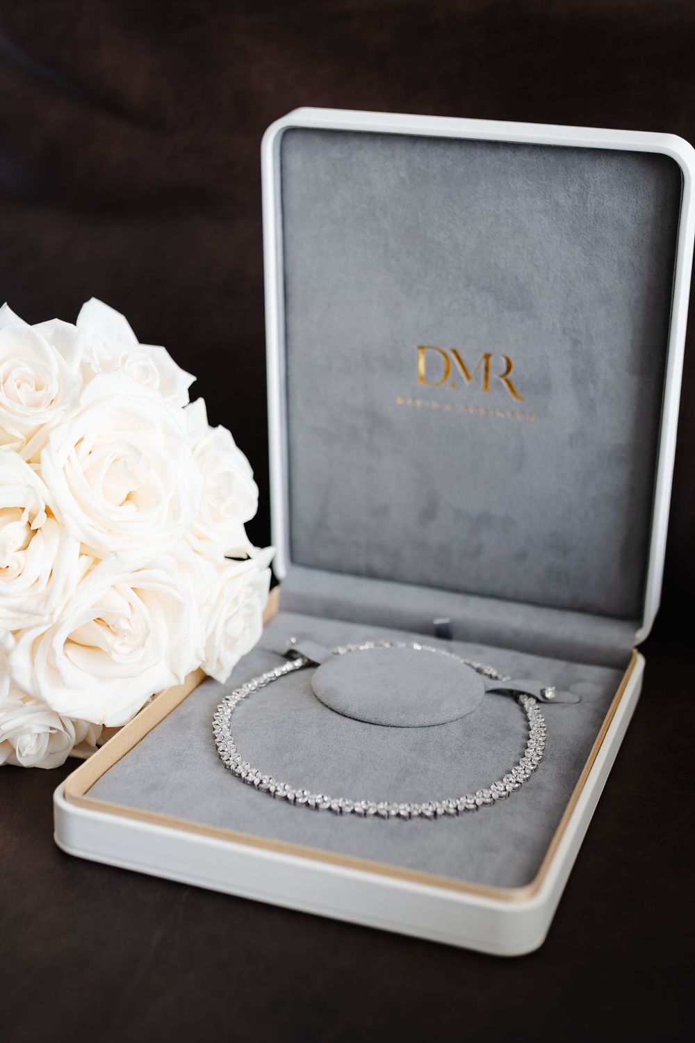 Diamond tennis necklace in a gray velvet DNR jewelry box beside a bouquet of white roses.