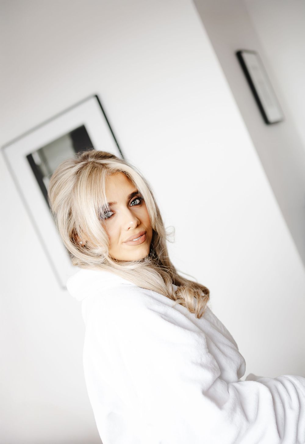 Beautiful blonde bride in a white robe smiling over her shoulder in a bright hotel room.