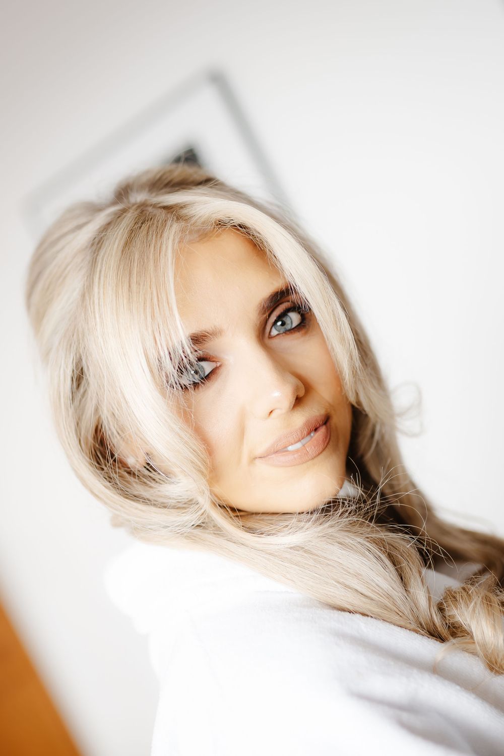 Blonde woman with blue eyes and long flowing hair poses for a close-up portrait in a bright white room.