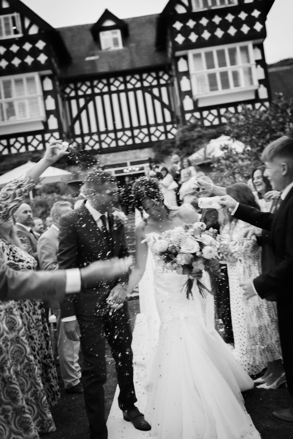 Wedding guests spray champagne in celebration outside a Tudor-style building.