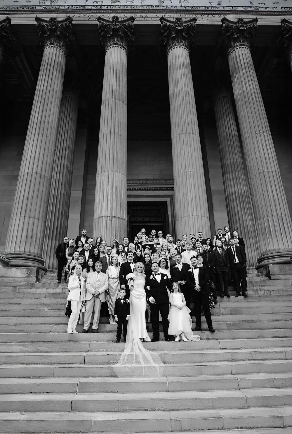 Black and white wedding photo of bride, groom, and guests on grand steps of a neoclassical building with tall columns.