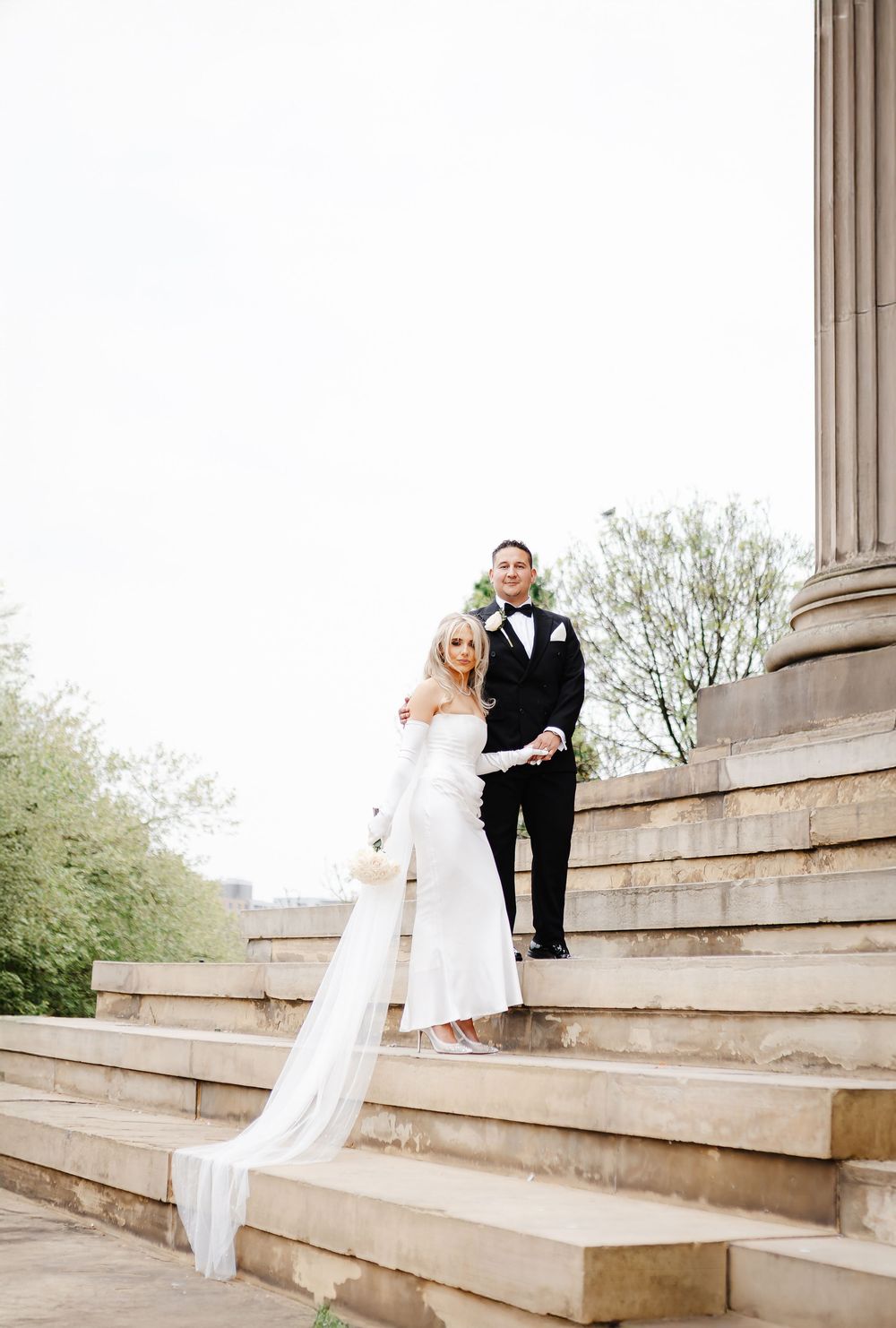Bride and groom posing on stone steps beside a classical column on their wedding day.