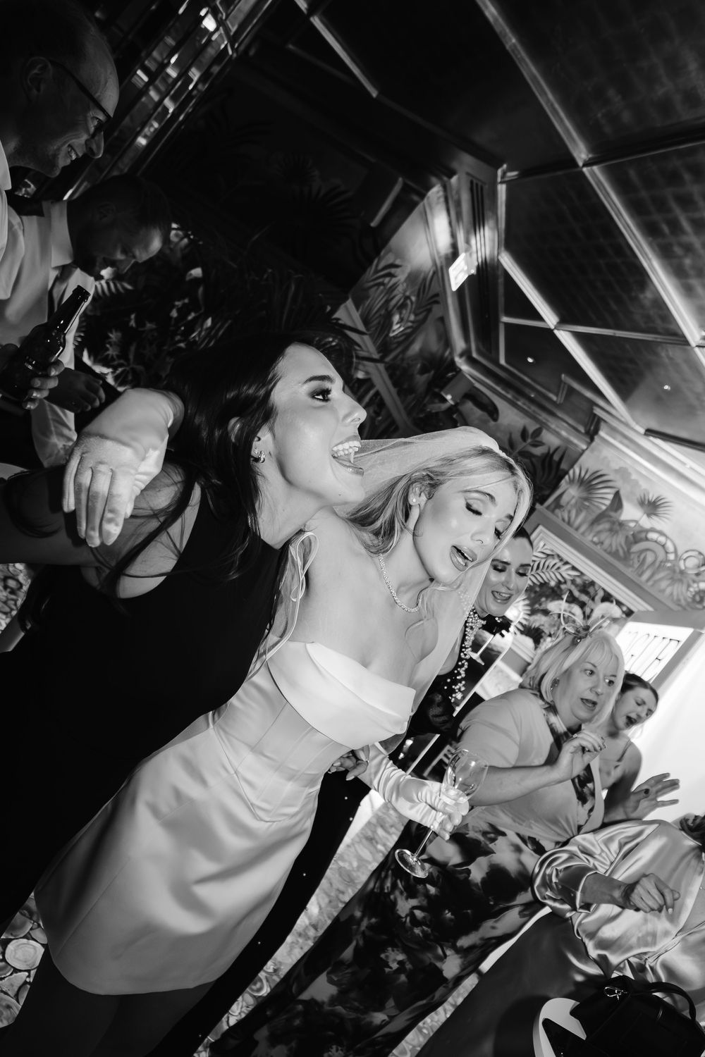 Black and white photo of two women dancing and laughing at a lively party or wedding reception event.
