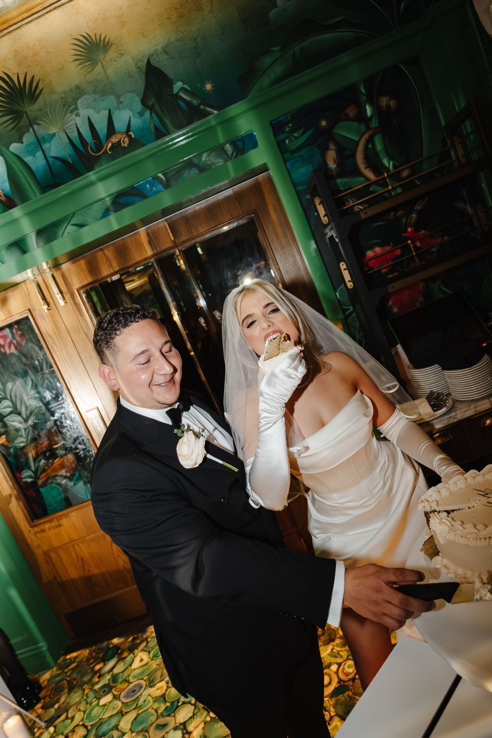 Bride and groom laughing during wedding cake cutting in elegant green-decorated venue, bride in off-shoulder gown with veil.