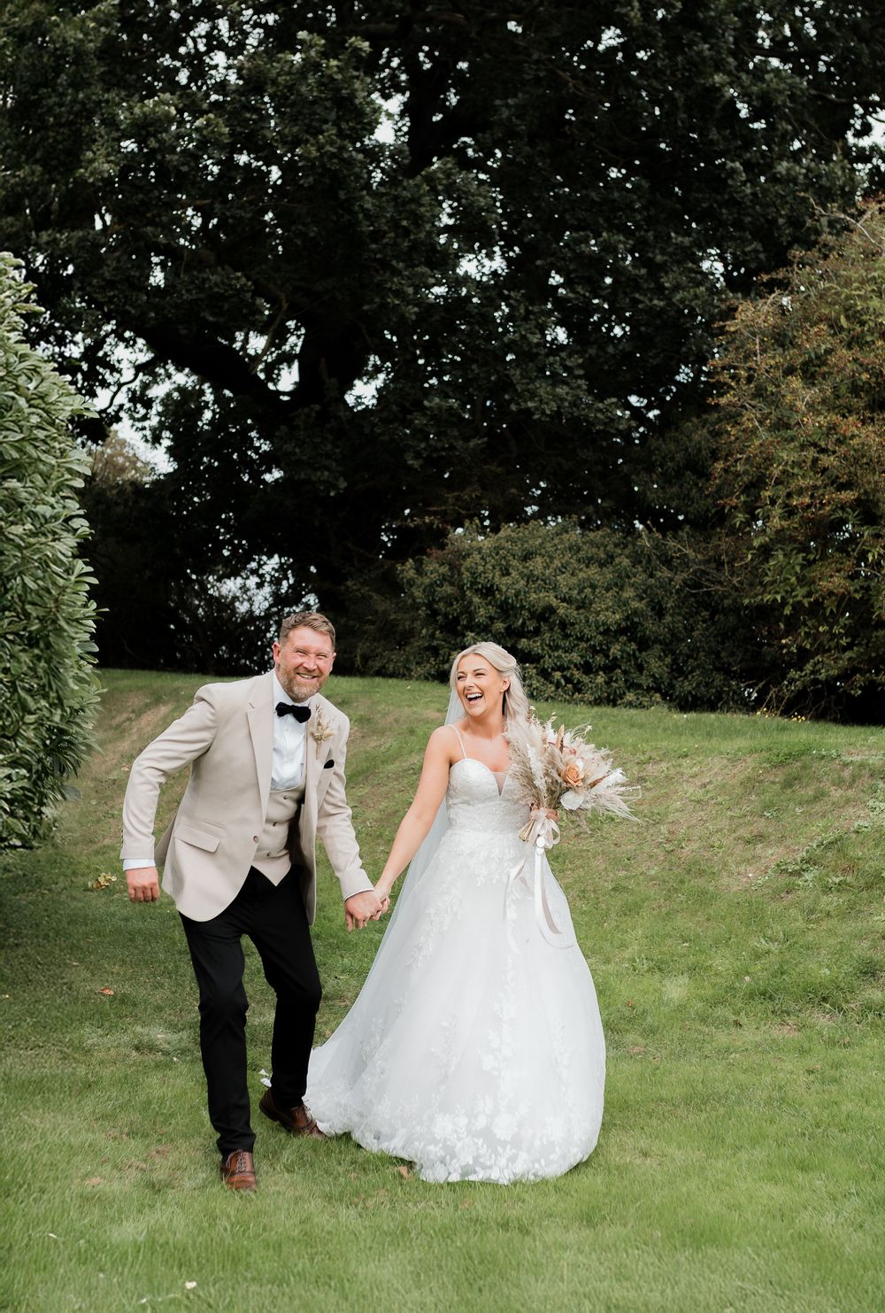 Laughing bride in white floral gown and groom in beige suit hold hands walking through lush garden on wedding day.