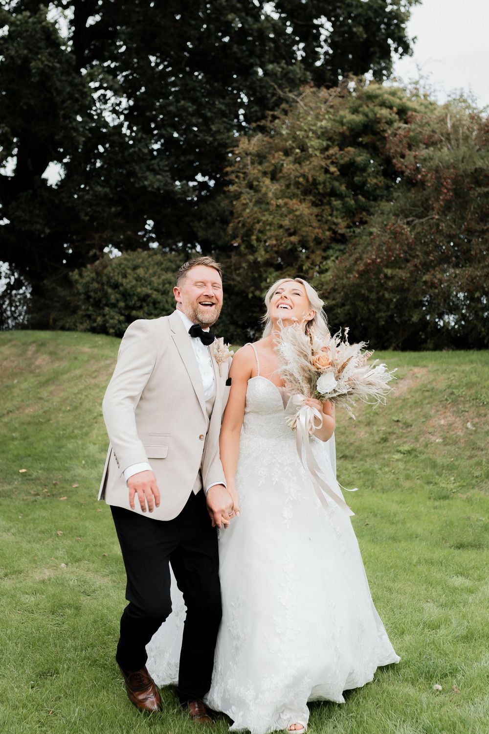 Laughing bride and groom walking outdoors on grass holding hands with boho wedding bouquet.