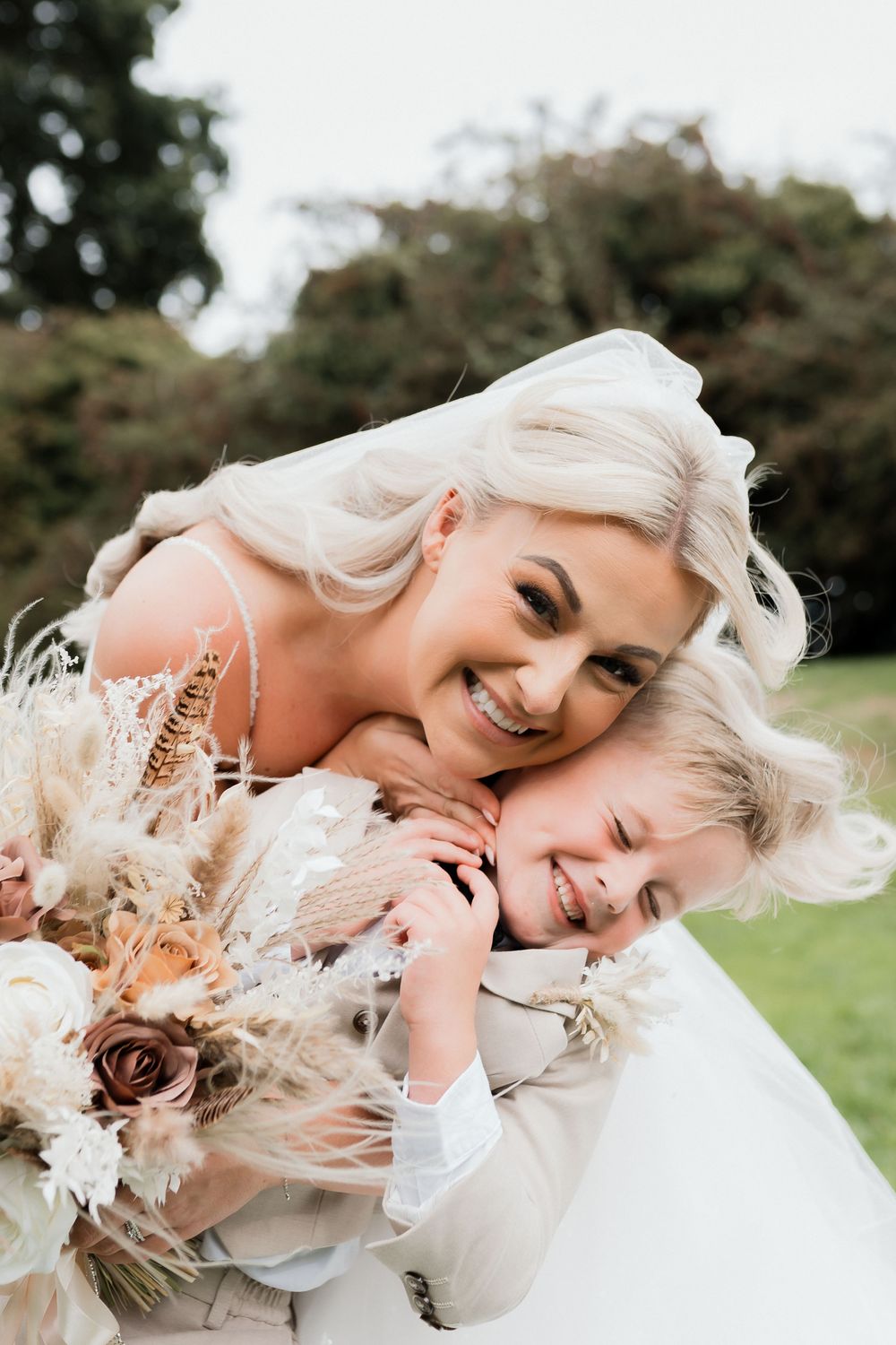 Smiling bride in white gown hugging a young child, holding a boho bouquet with dried flowers outdoors.