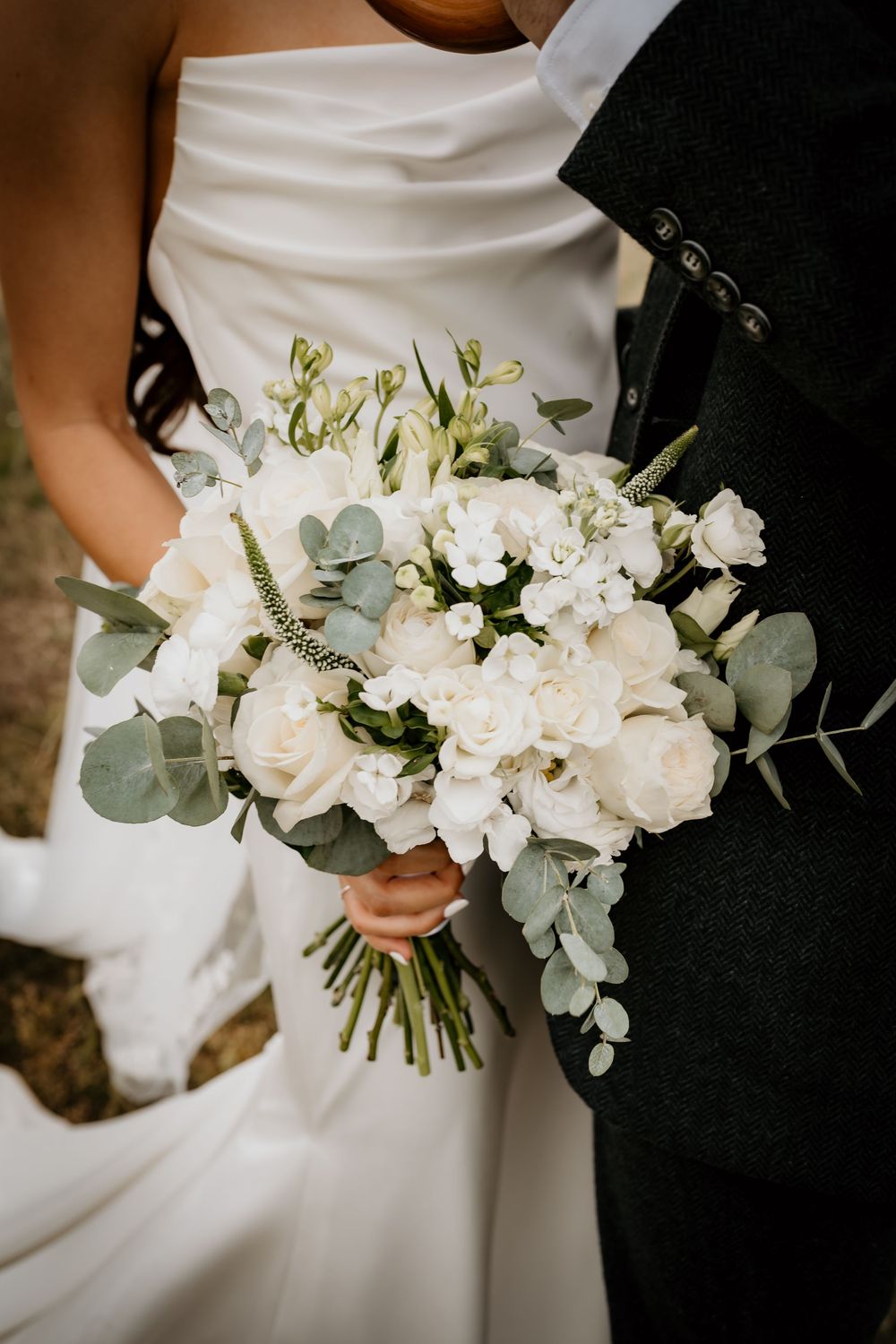 A beautiful white wedding bouquet with roses, eucalyptus leaves and delicate flowers held against a white dress.
