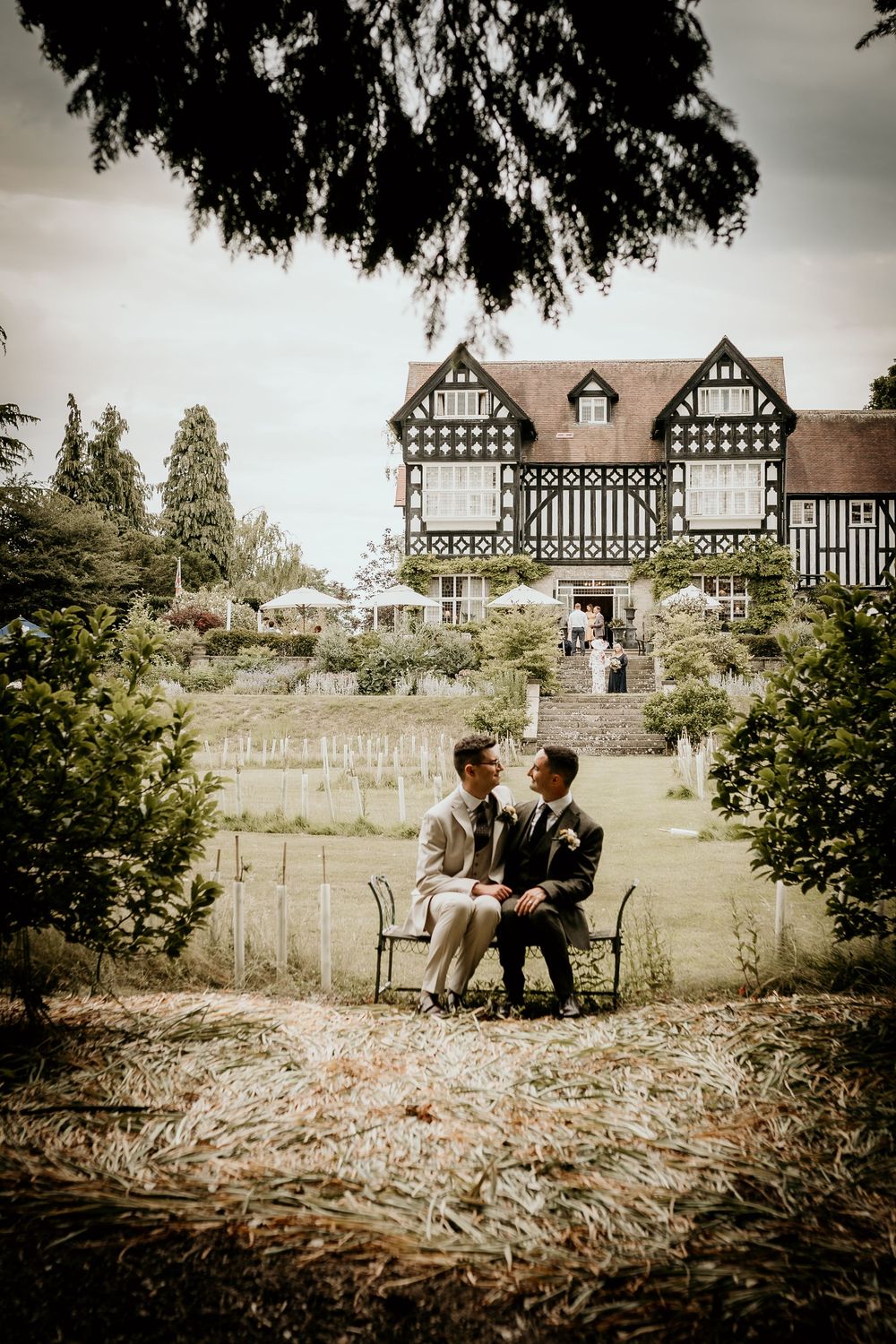 Couple sitting together on rustic bench with Tudor-style houses in background creating romantic countryside scene.