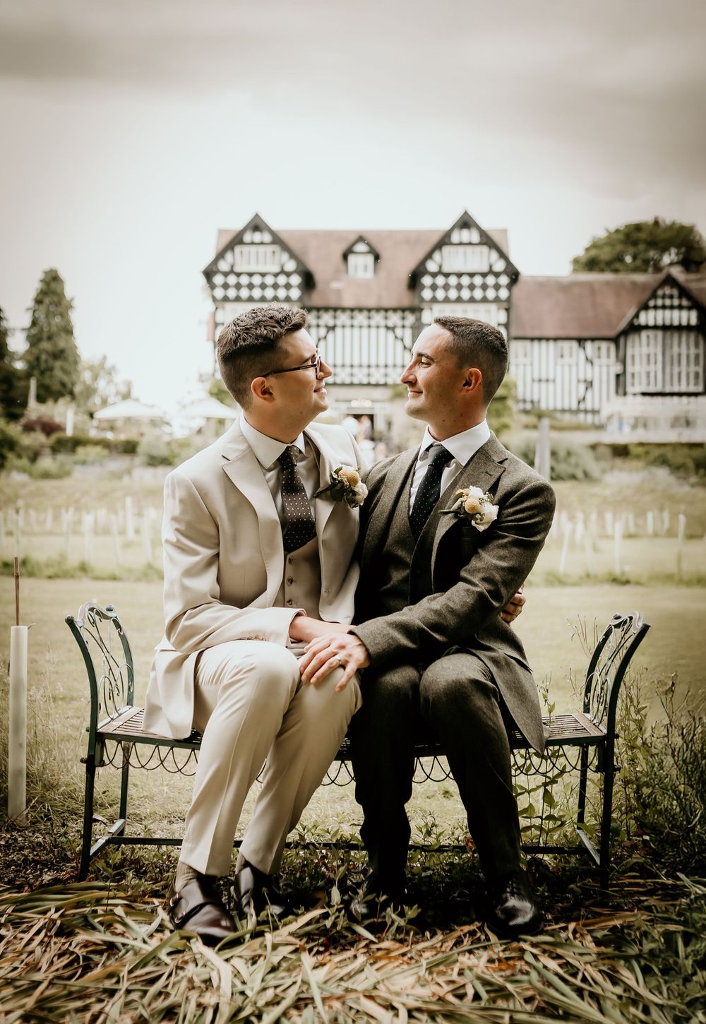 Two people sharing intimate moment on outdoor bench with traditional Tudor architecture in pastoral setting.