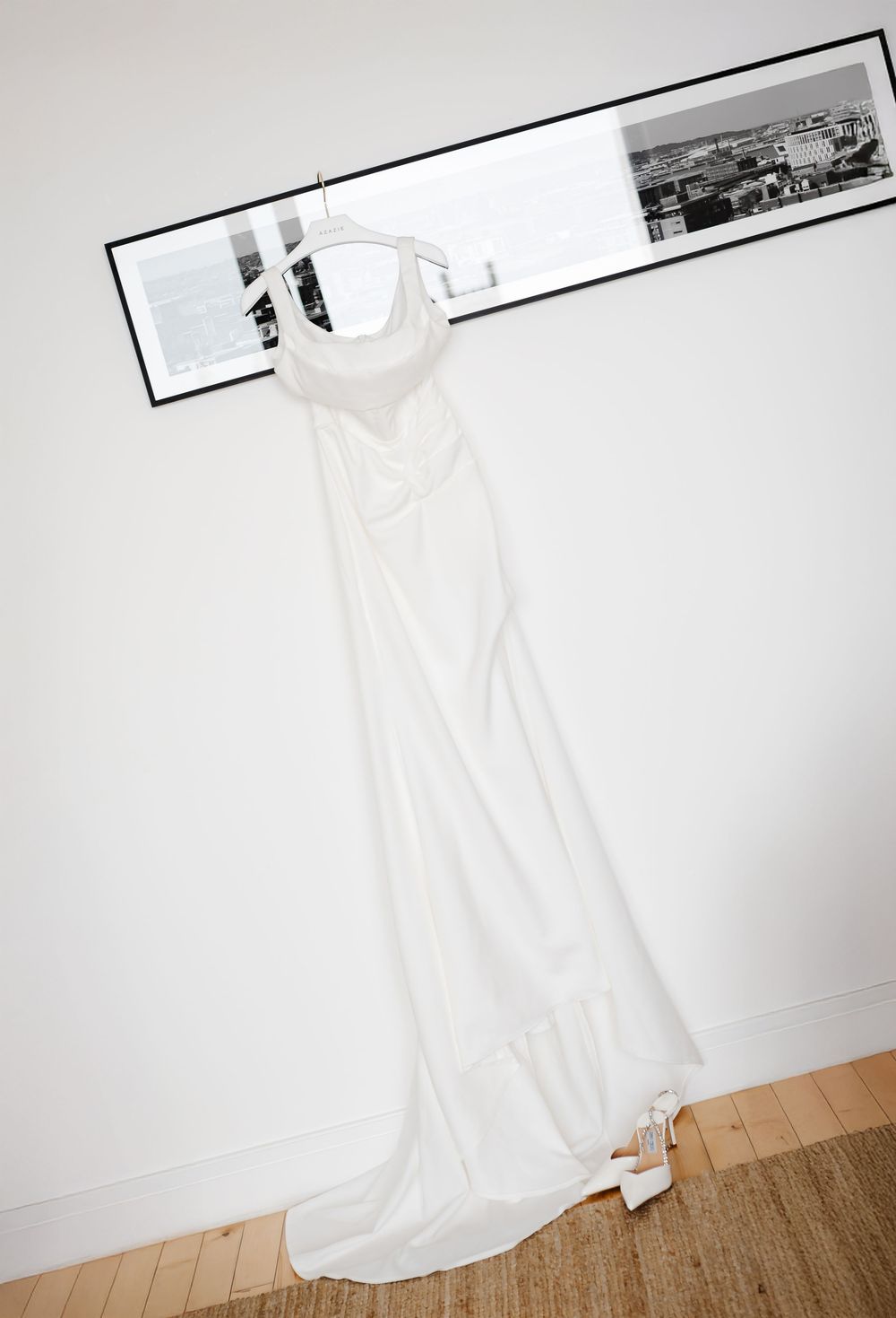 Elegant white wedding dress hanging on a hanger against a white wall with black and white framed art.