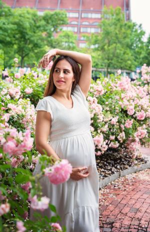 A woman in a white summer dress poses gracefully among blooming pink roses in an urban garden setting.