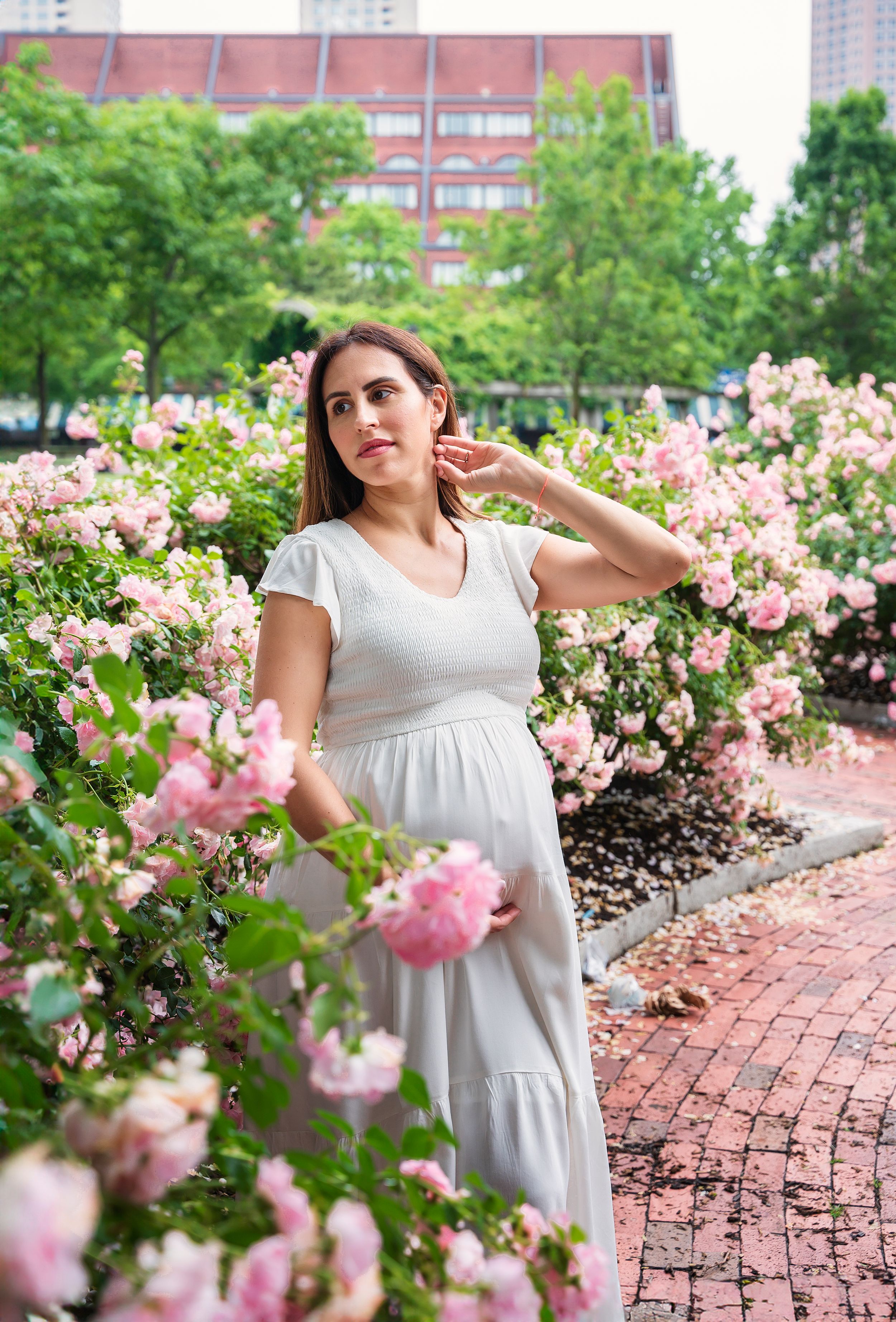 A white dress stands elegantly among blooming pink roses along a brick pathway in an urban garden setting.