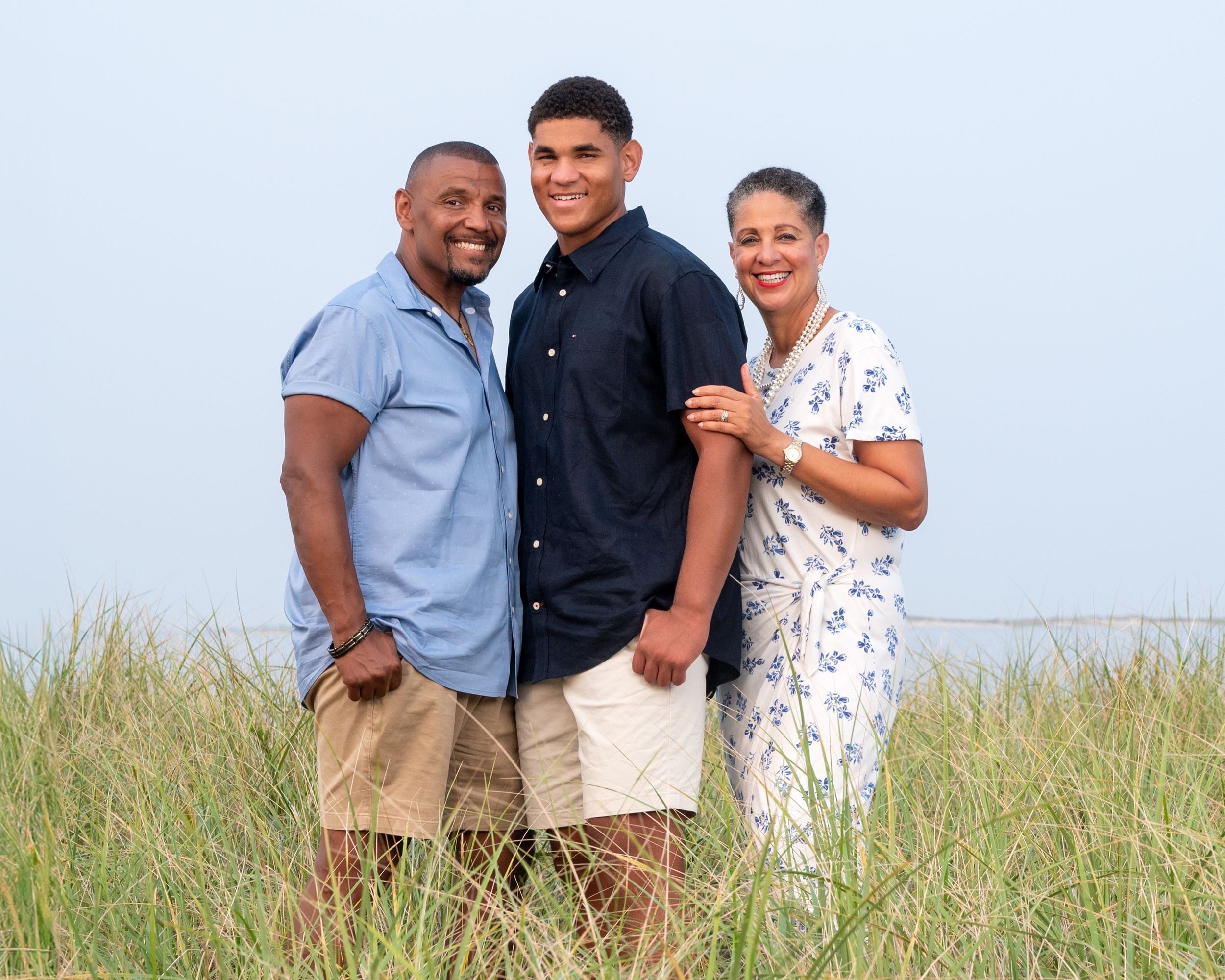 Three people stand together smiling in a grassy field with tall grass and blue sky in the background.