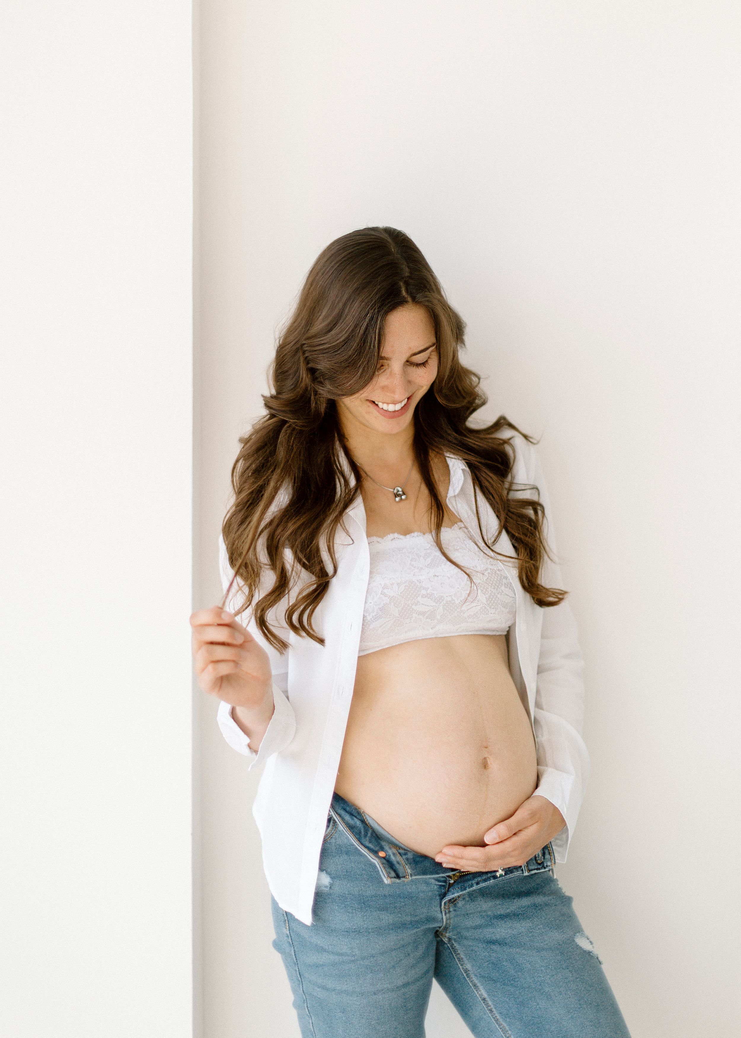 A pregnant woman in a white button-up shirt and jeans stands against a white wall showing her bare belly.