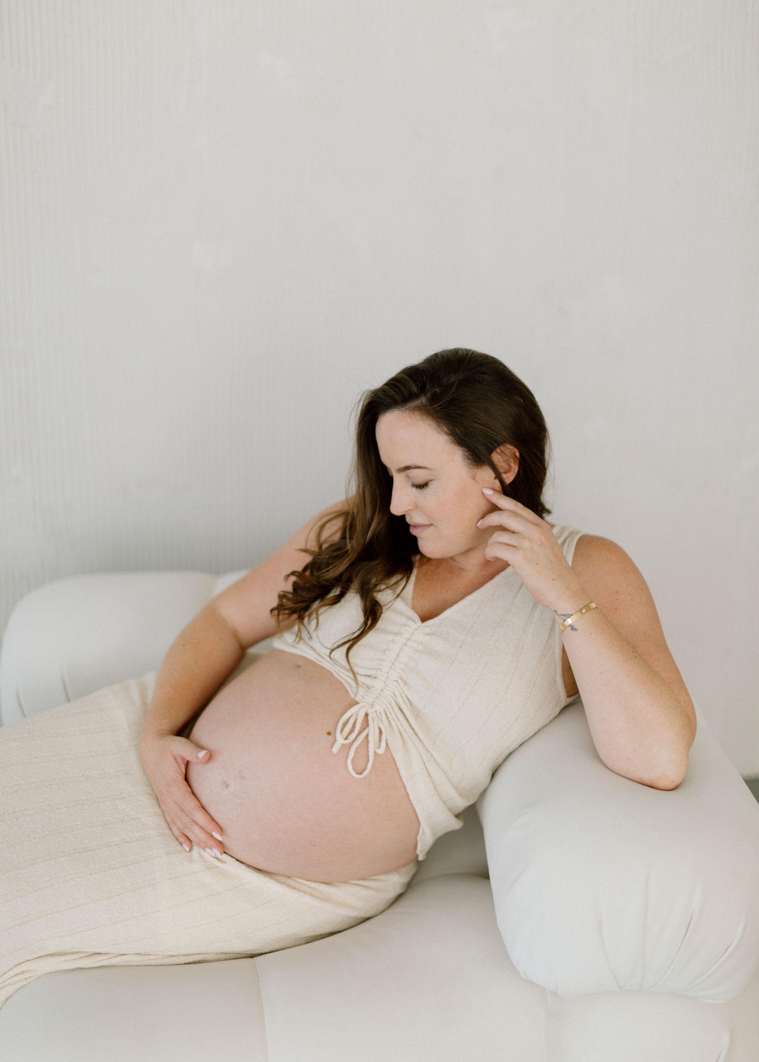 A series of minimalist maternity photos showing a pregnant person in a cream dress lying on white bedding.
