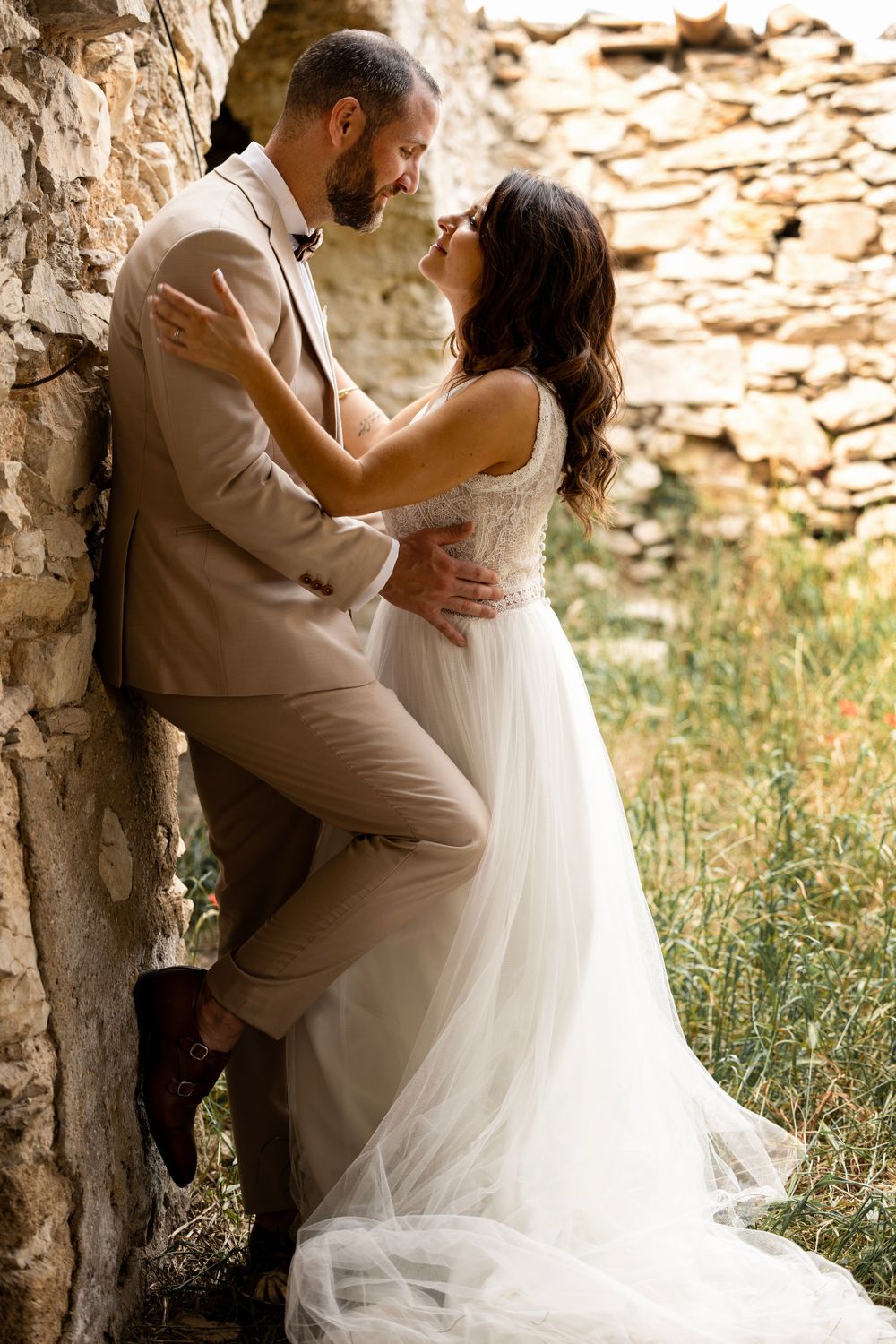 A romantic wedding couple embraces against a stone wall backdrop, with the bride in a flowing white dress and groom in tan suit.