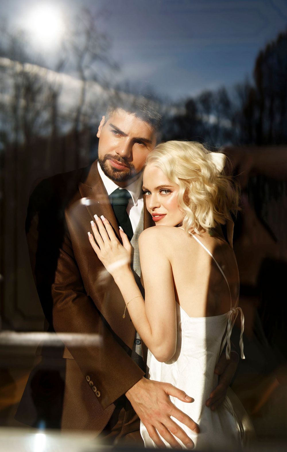 A romantic wedding portrait captured through glass with dramatic backlighting and elegant formal attire.