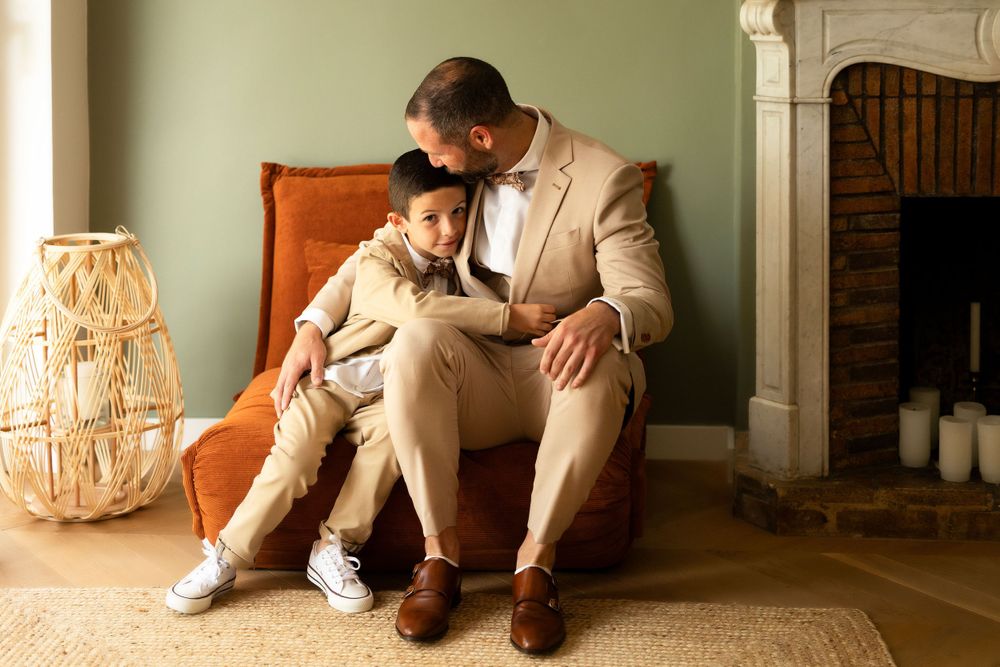 A parent and child in matching beige suits sit together on an orange armchair near a decorative wicker lamp.