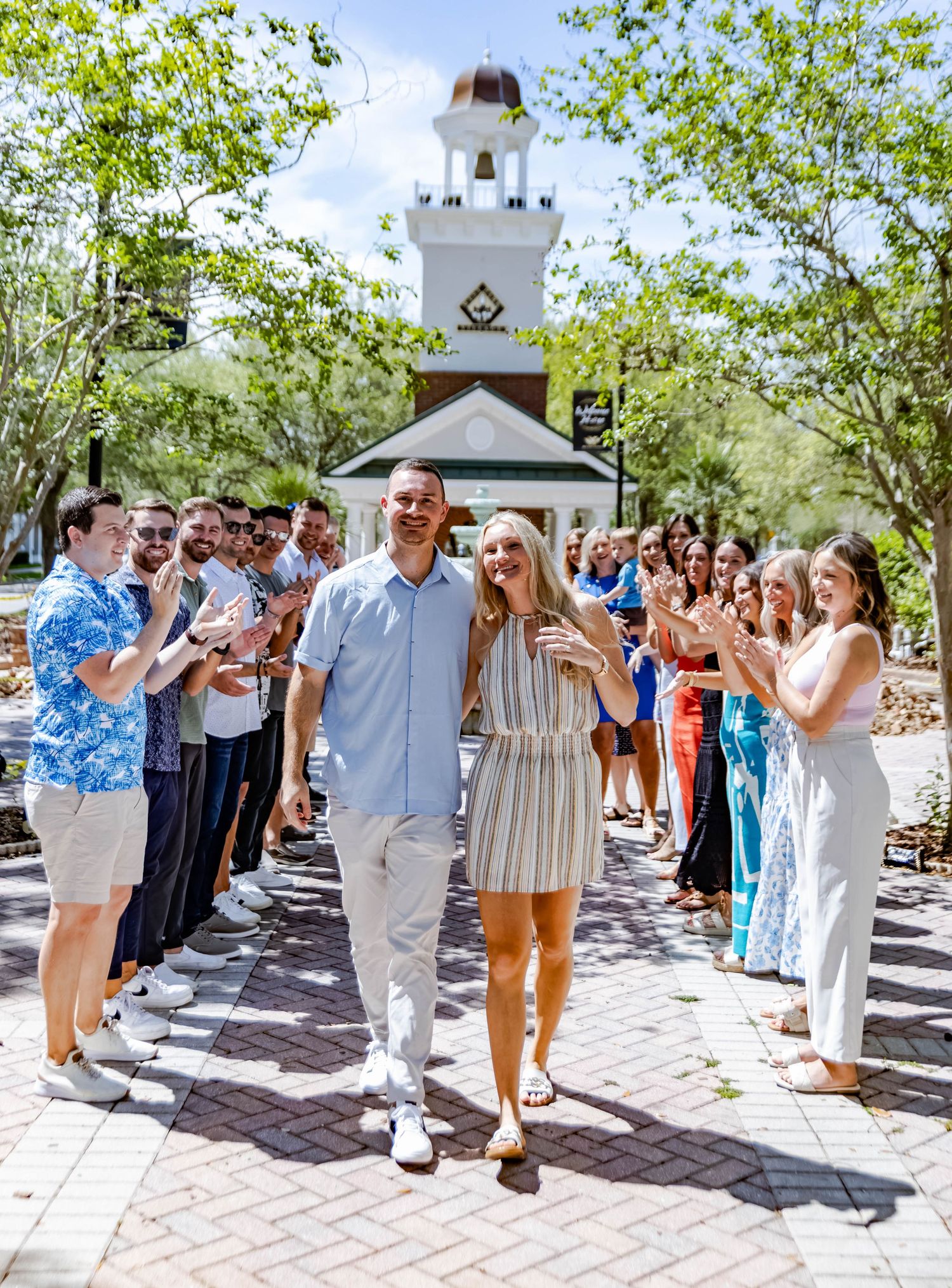 A couple walks down a brick pathway lined with cheering onlookers and greenery on a sunny day.