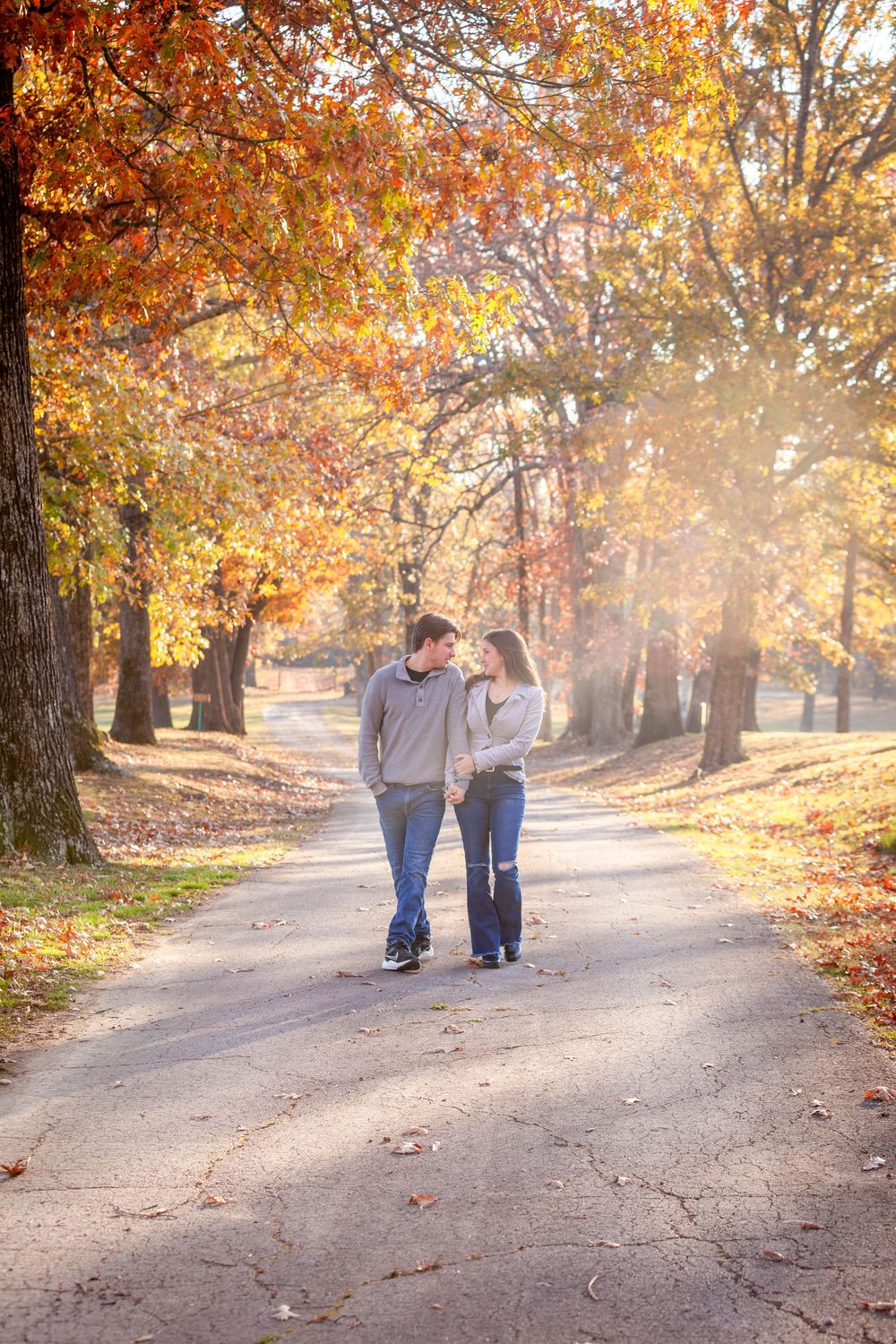 engaged couple holding hands walking down a tree-lined gravel road in sunset light beneath the fall leaves and color in Batesville, Arkansas