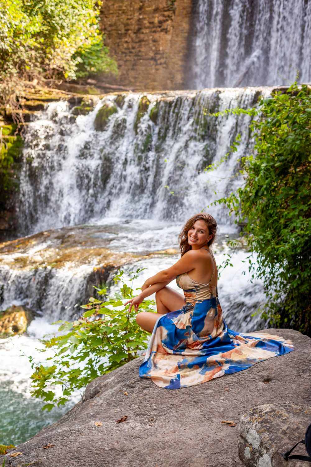 senior girl squatted on a rock in long blue dress in front of a waterfall below Mirror Lake Dam at Blanchard Springs in Fifty-Six, Arkansas
