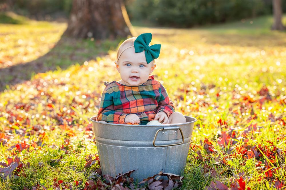 Toddler girl in fall colored outfit with bow headband sitting in metal bucket in the grass and leaves in the evening in Batesville, Arkansas