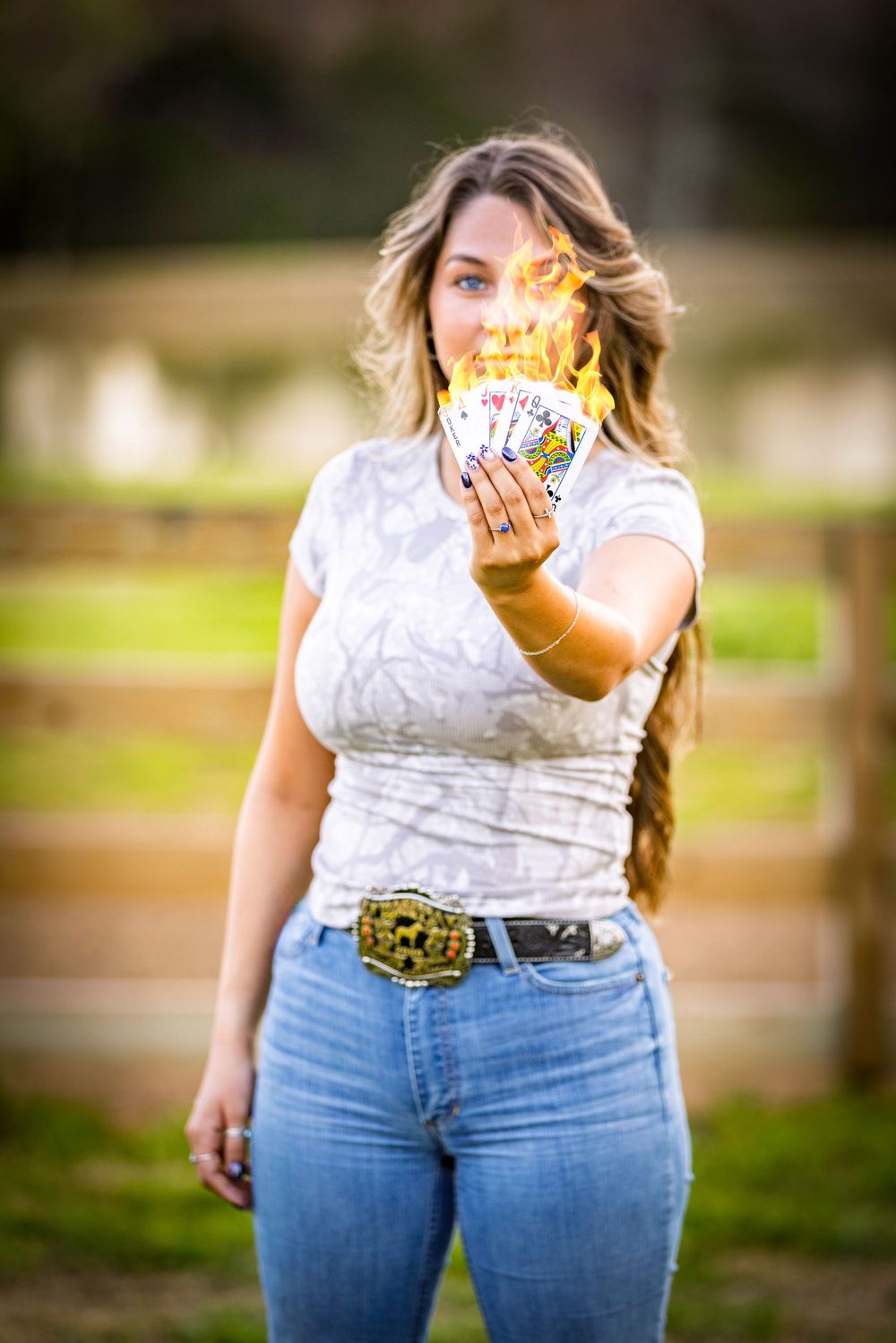 senior girl holding out a handful of playing cards in front of her that are on fire in front of a fence and pond outside Searcy, Arkansas