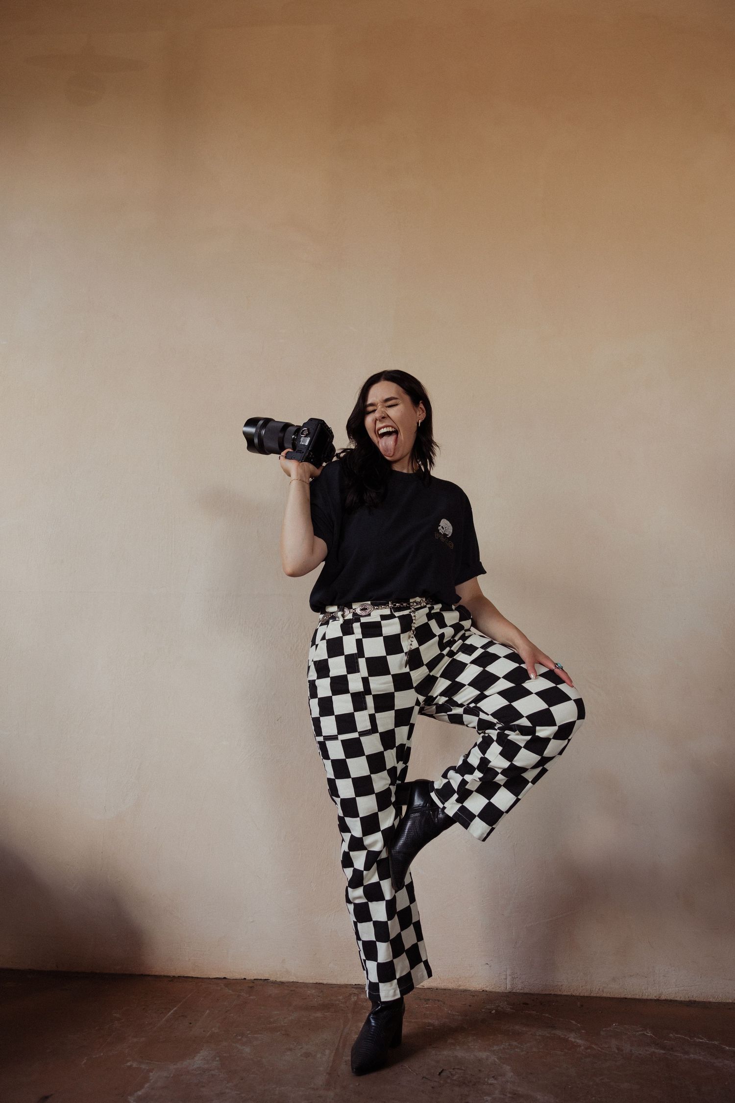 A photographer wearing checkered pants and black shirt poses energetically with a camera against a beige wall.