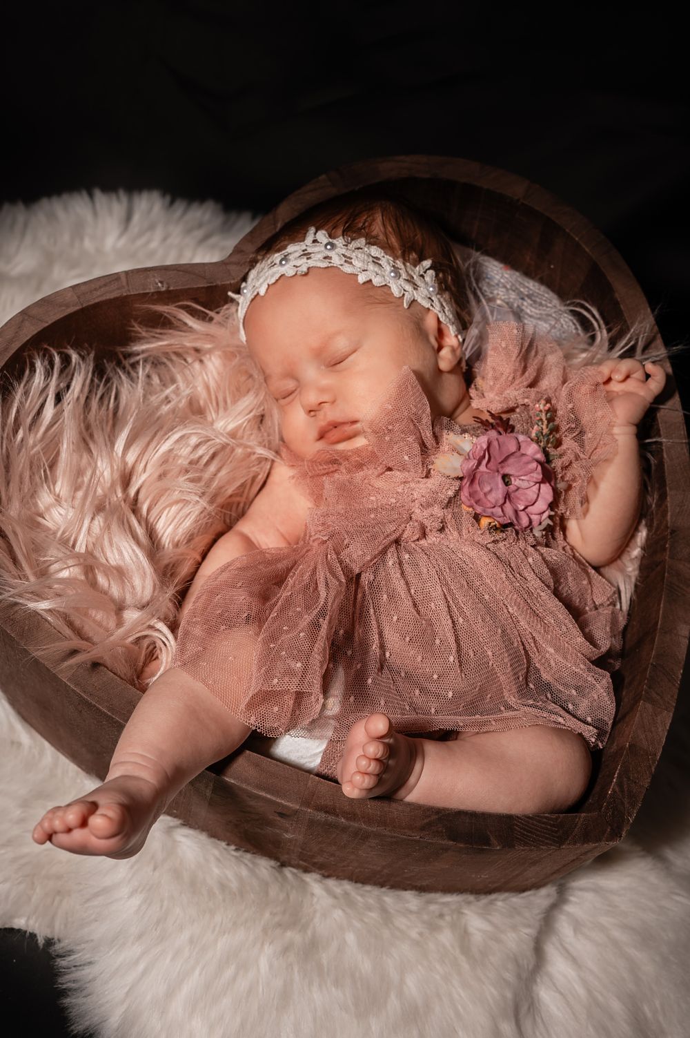 A sleeping baby in pink tulle dress lies peacefully in a wooden bowl on white fur background.