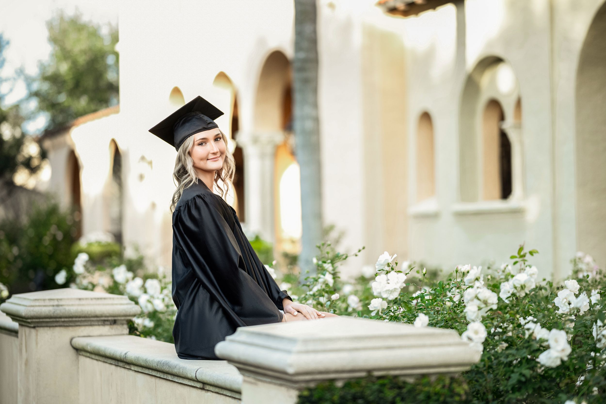 Graduate in cap and gown sits on stone ledge near white building with arches.