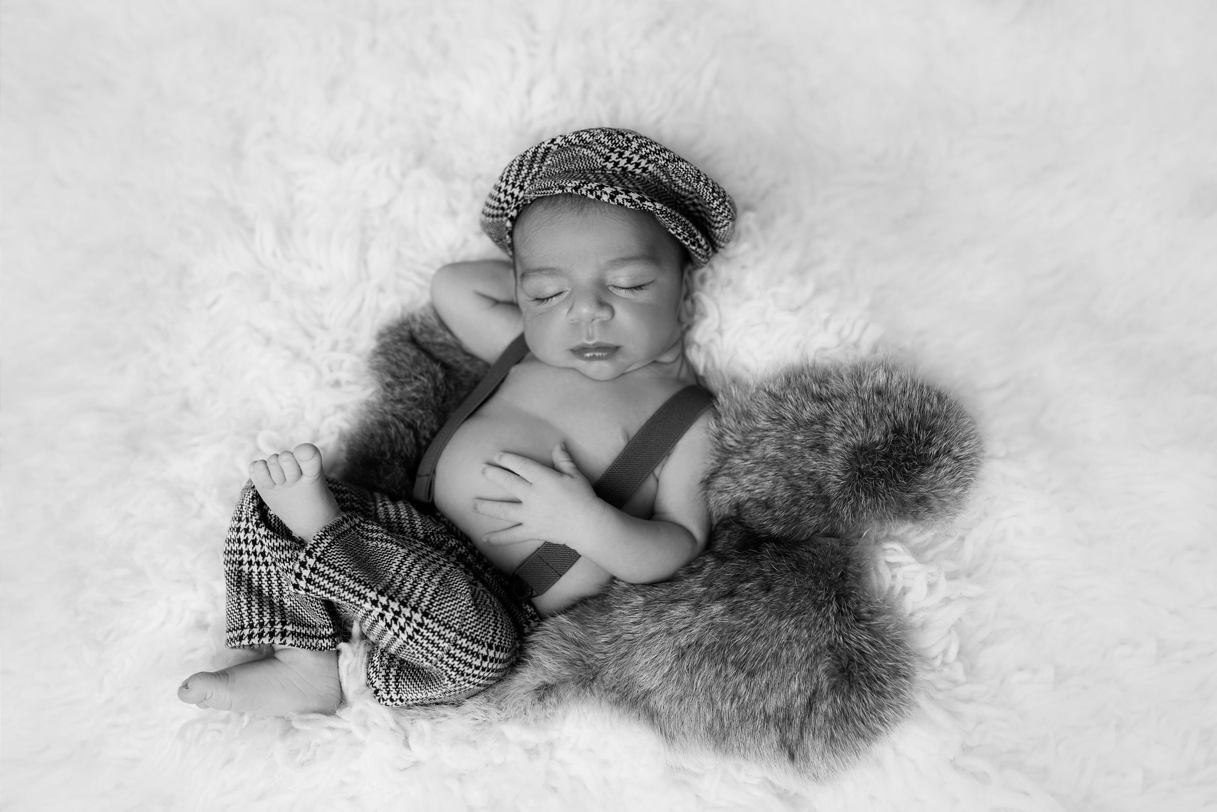 Sleeping newborn in newsboy cap surrounded by soft fur on white background.
