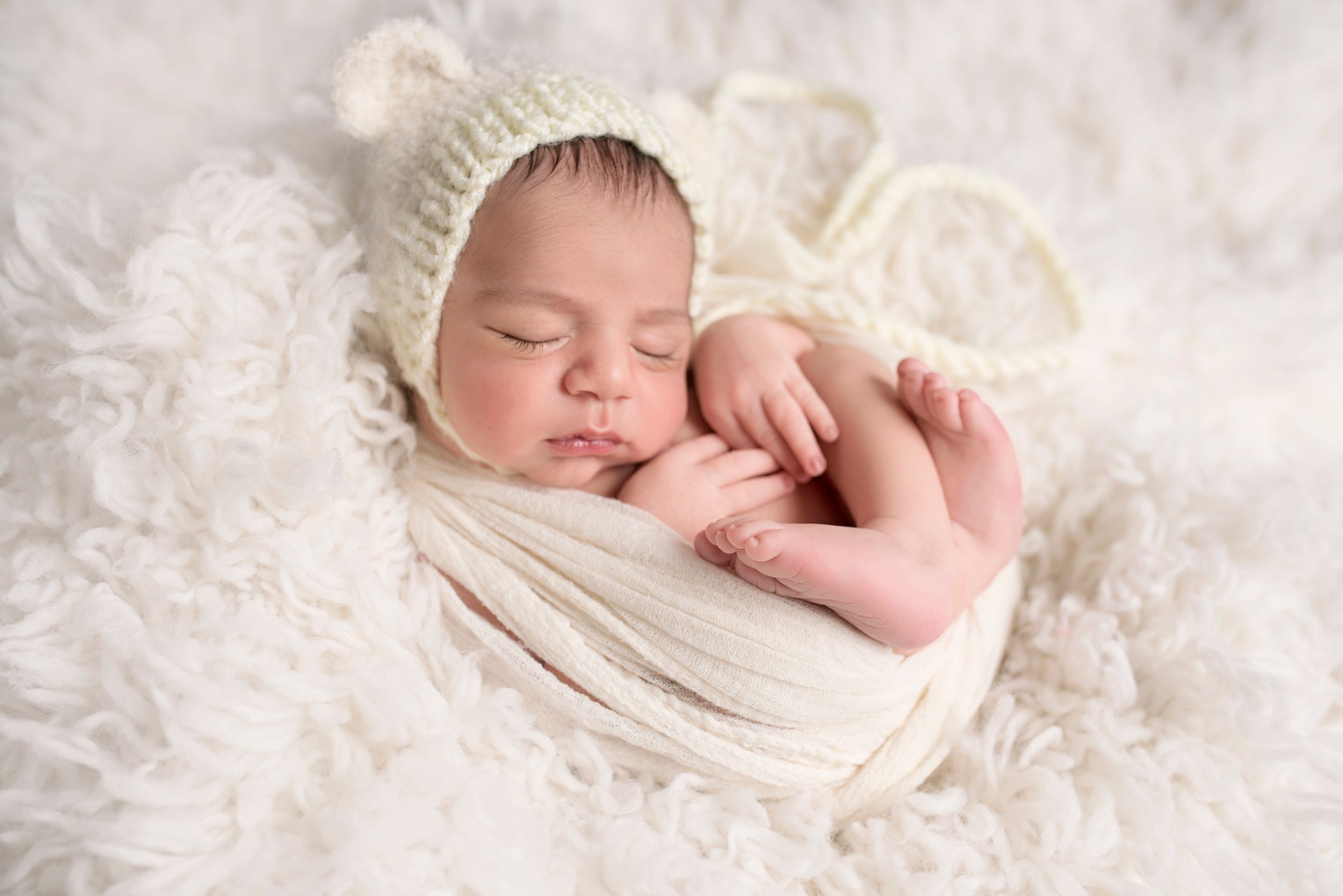 Peaceful sleeping newborn wrapped in white wearing knit bear bonnet on fluffy backdrop.