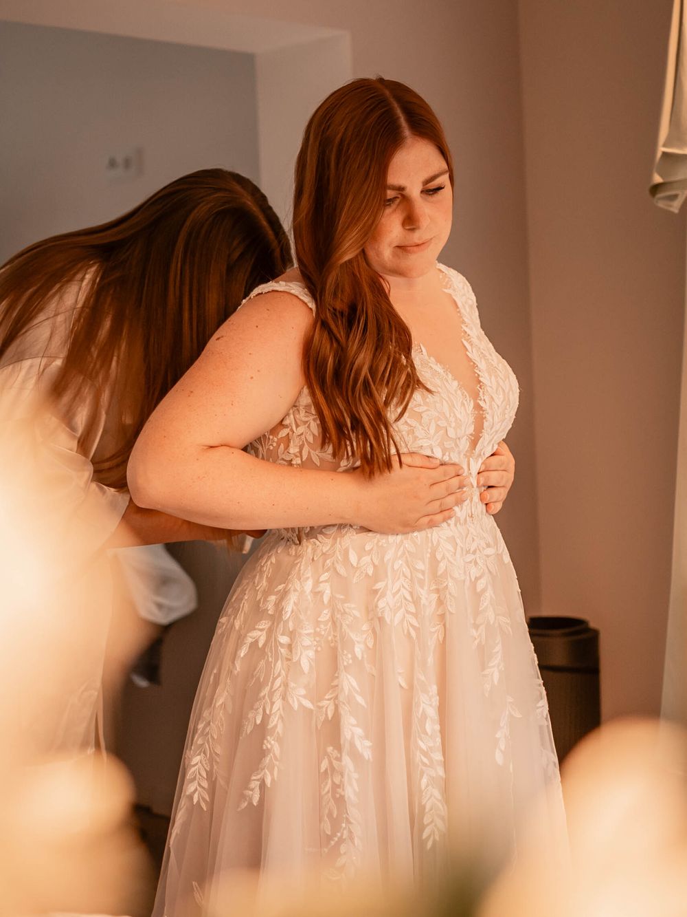 A woman wearing a delicate white lace wedding dress with floral details in a softly lit room during preparations.