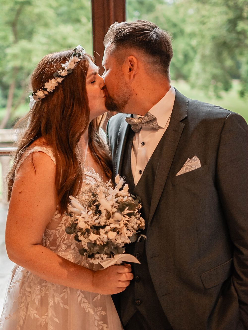 A romantic wedding moment showing a happy couple sharing a kiss while the bride holds a delicate dried floral bouquet.