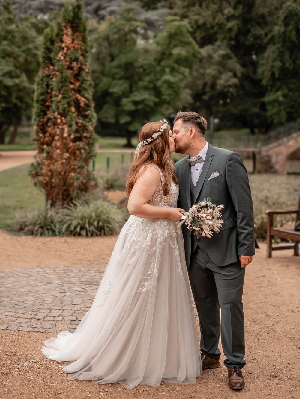 Romantic outdoor wedding couple in embrace on cobblestone path with bride wearing flowy lace gown and holding dried bouquet.