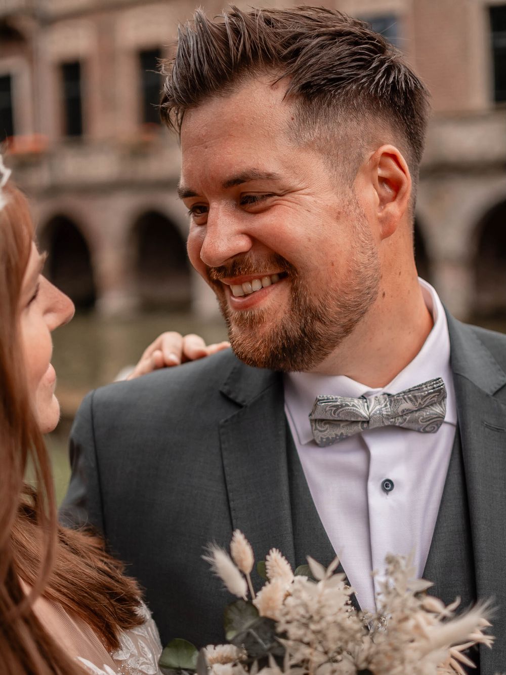A romantic wedding moment featuring a groom in a grey suit and silver bowtie smiling at his partner.