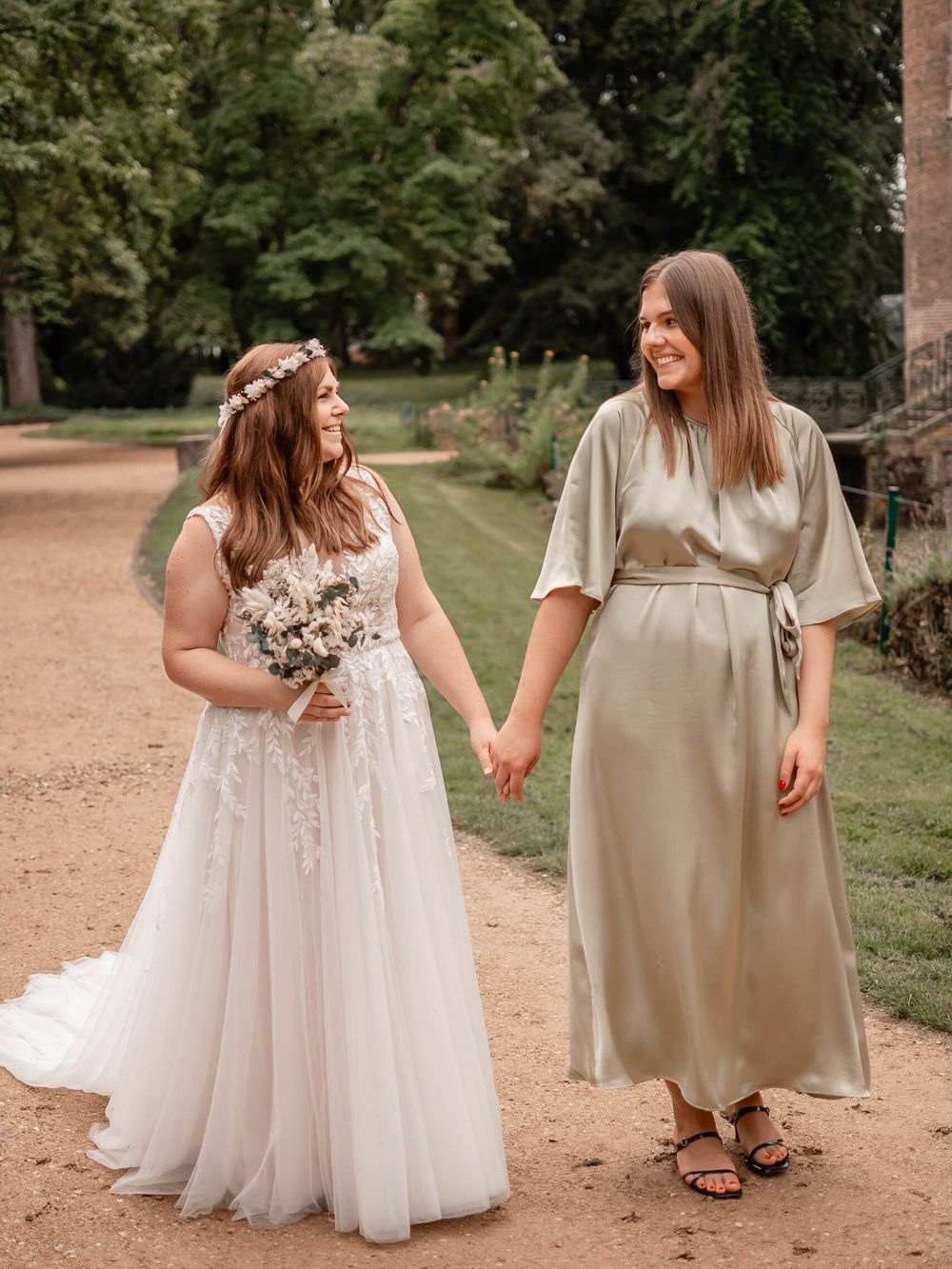 Two people wearing matching champagne and white dresses stand together on a dirt path in a garden setting.