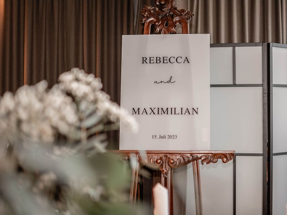 Minimalist wedding sign for Rebecca and Maximilian displayed in an ornate copper frame with soft floral elements.