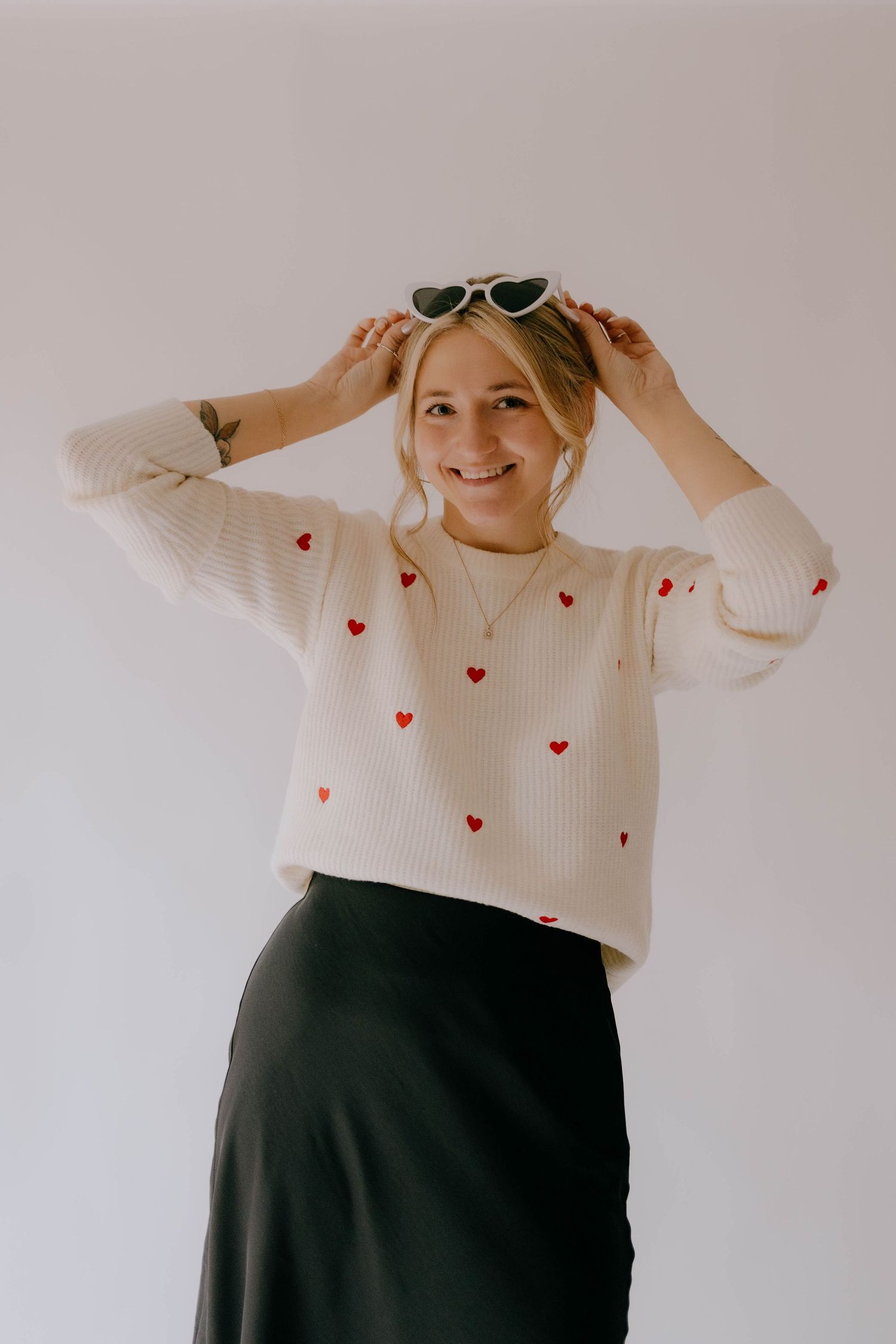 Playful pose in cream sweater with heart pattern and black skirt against neutral background.