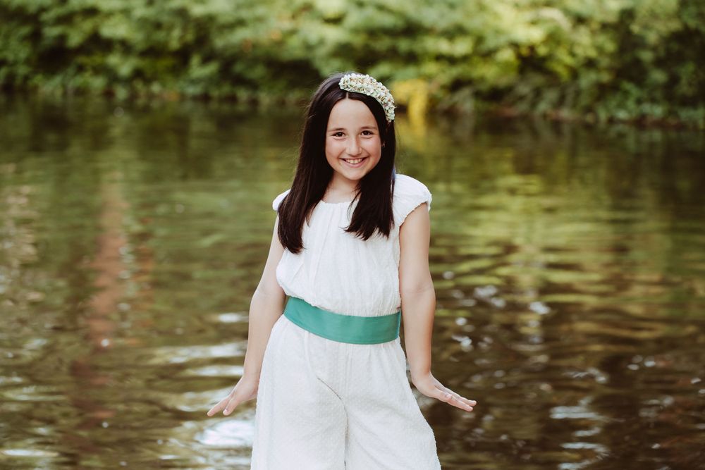 Person in white dress with green sash standing by reflective pond in nature setting.