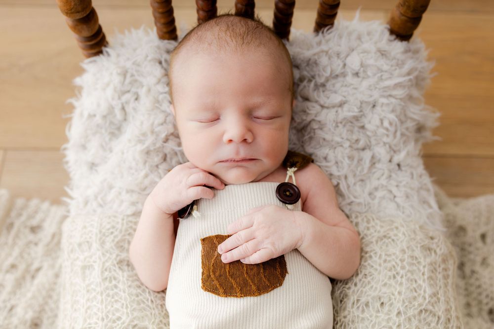 Peaceful newborn baby sleeping in a wooden chair on a knit blanket, wearing a cute bear outfit.