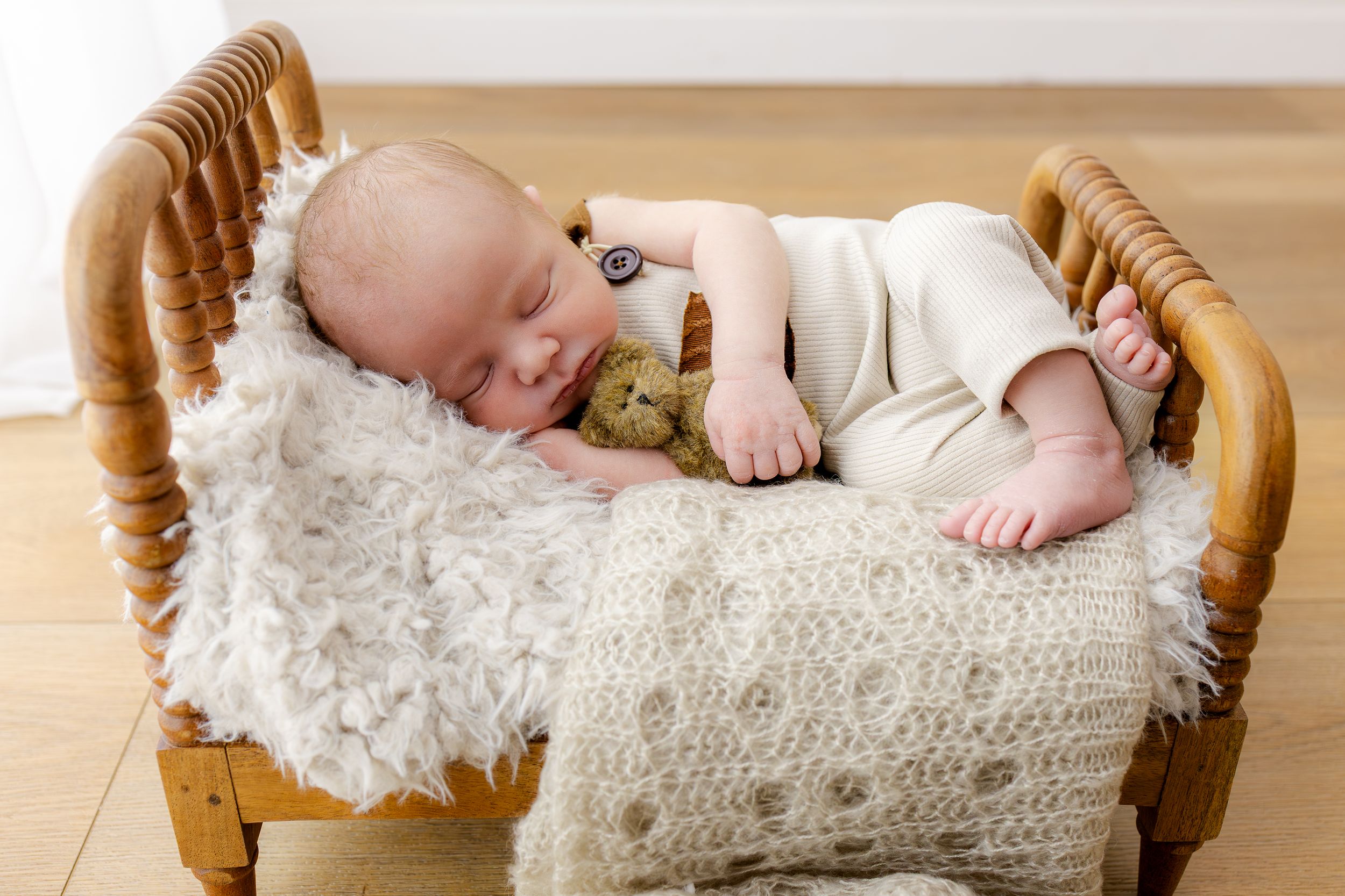 Sleeping newborn baby in tiny wooden bed with teddy bear and fluffy white blanket.