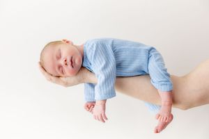 Newborn baby sleeping peacefully in blue onesie cradled on an adult hand against white background.