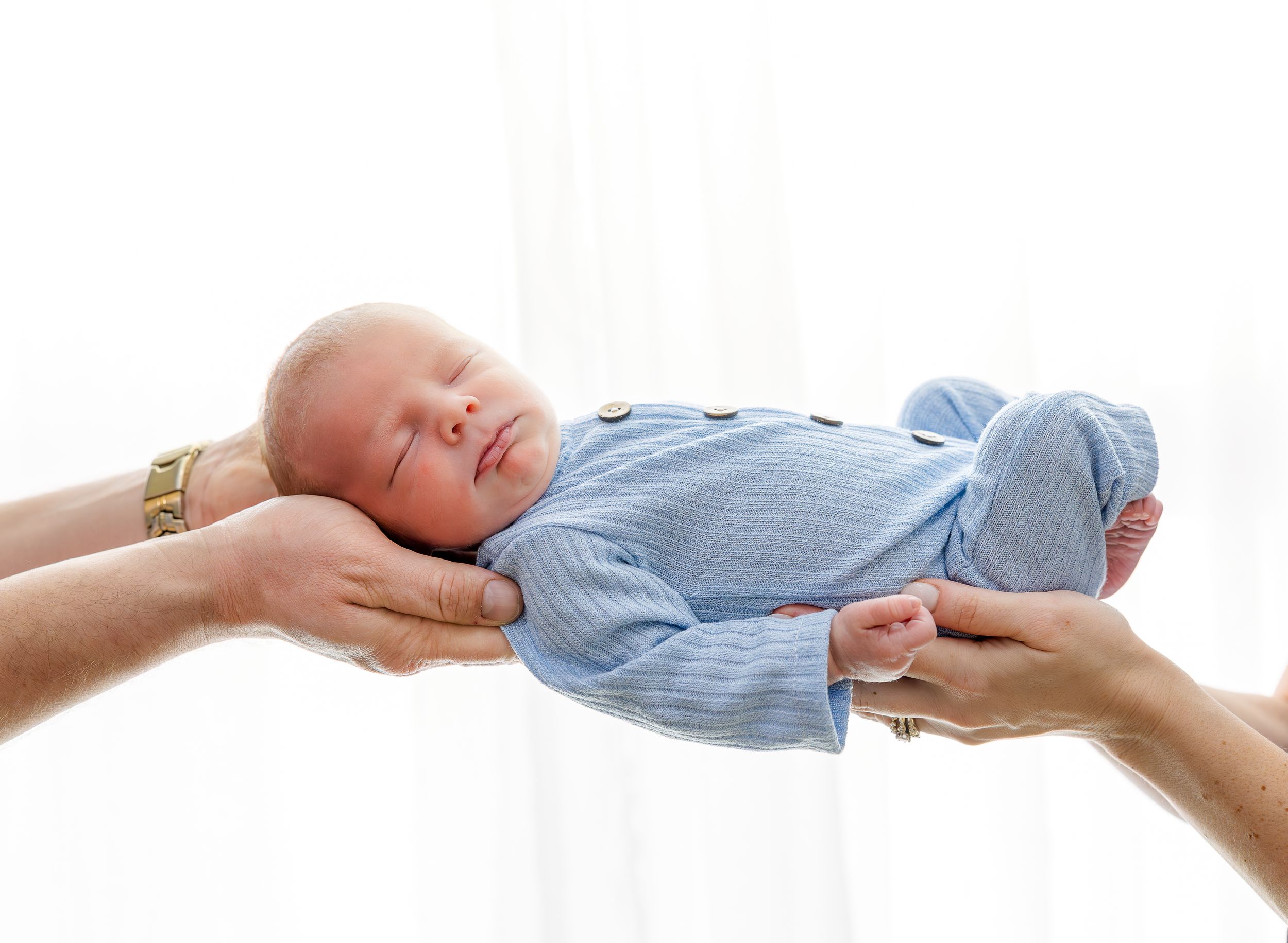 Newborn baby in blue onesie sleeping peacefully while being gently held by two pairs of adult hands.