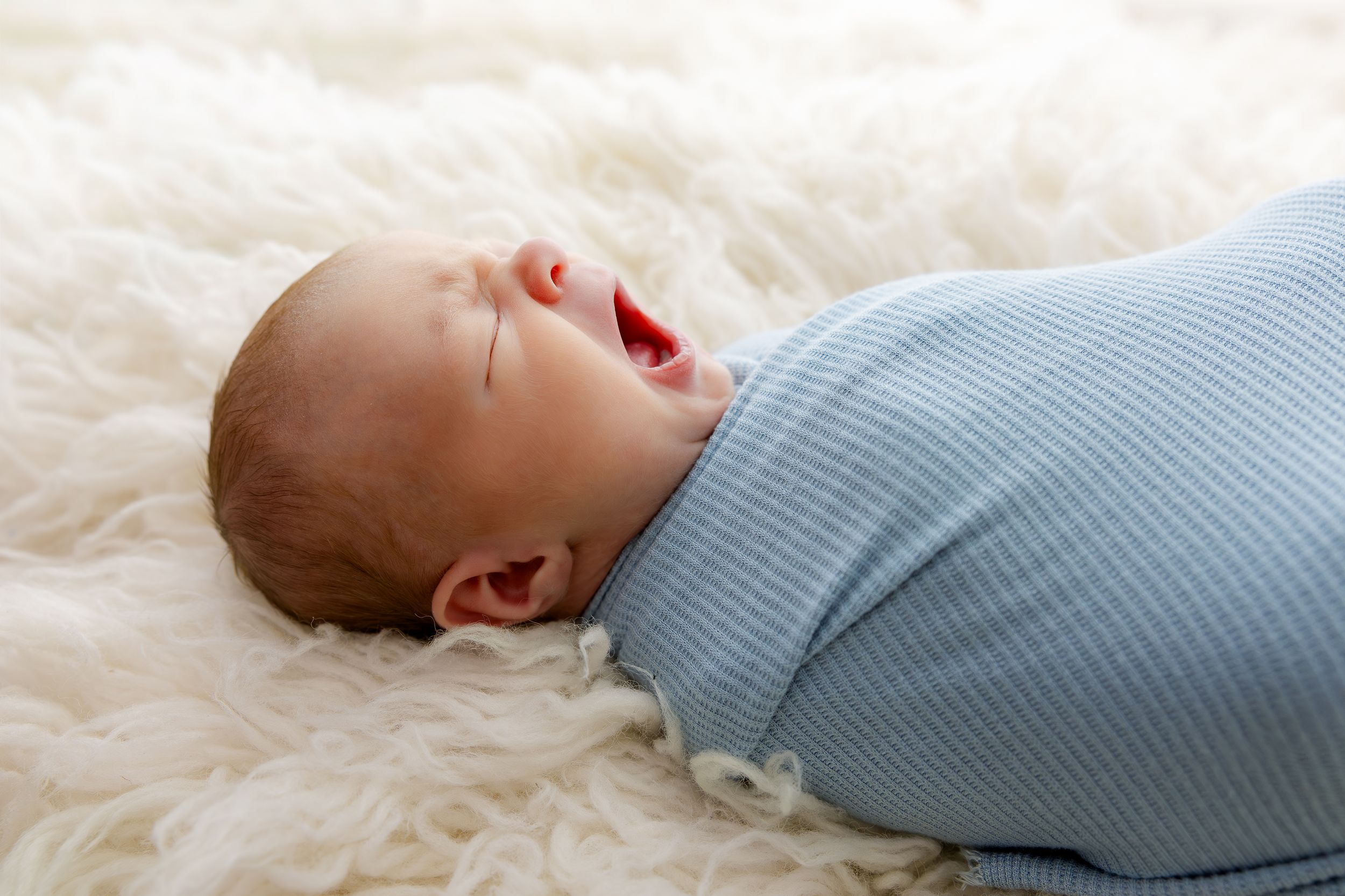Newborn baby swaddled in a blue striped wrap, yawning peacefully on a soft white fluffy blanket.