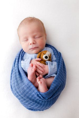 Sleeping newborn baby swaddled in blue wrap holding a small teddy bear on white background.