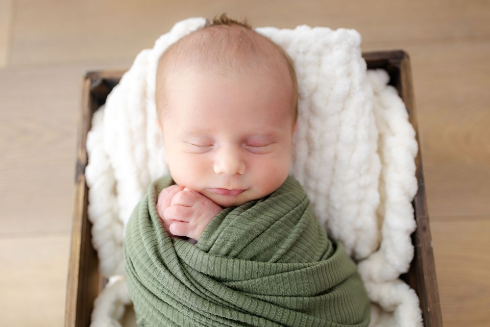 Sleeping newborn wrapped in sage green swaddle on white cushioning.
