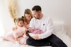 Family wearing white and pink outfits cuddles together on white bedding in a bright bedroom setting.