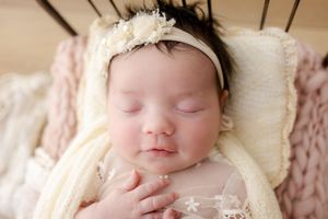 Sleeping newborn wearing white headband and lace wrap while resting on cream colored blanket.
